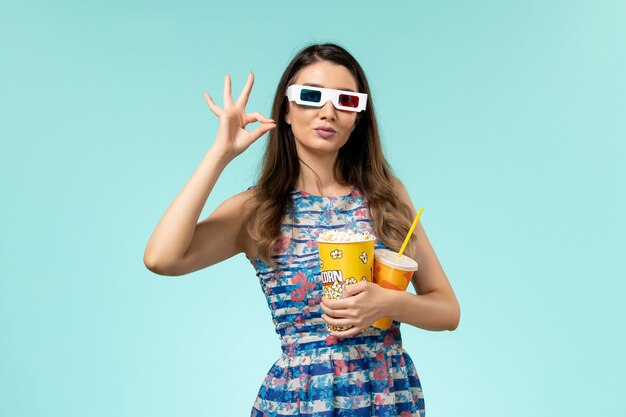 Vista frontale giovane femmina che tiene popcorn e bevanda in occhiali da sole d sulla superficie azzurra
