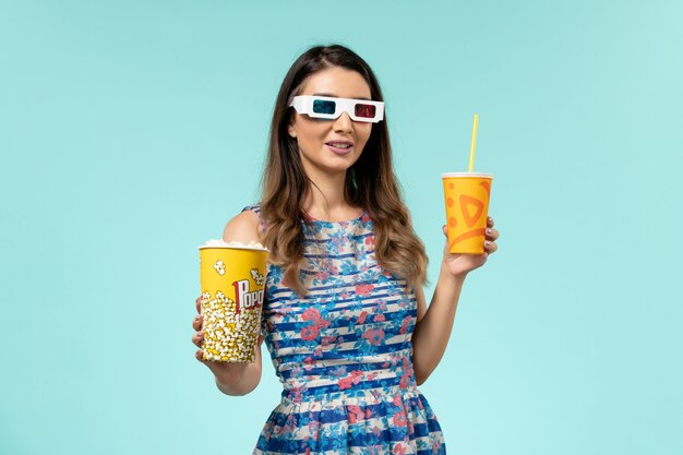 Vista frontale giovane femmina che tiene popcorn e bevanda in occhiali da sole d sulla superficie blu