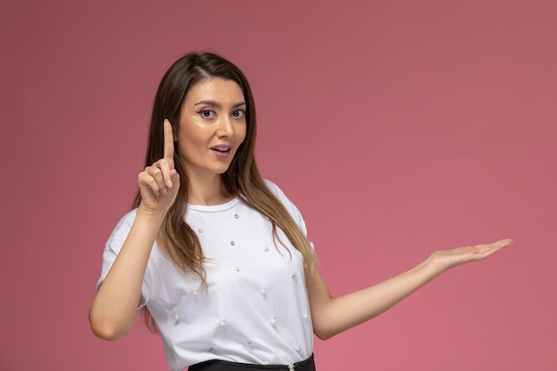 Vista frontale giovane femmina in camicia bianca che mostra il dito sul muro rosa, modello donna di colore