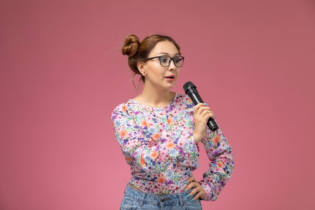 Vista frontale giovane femmina in camicia fiore progettato e blue jeans che tengono il microfono cercando di cantare sullo sfondo chiaro