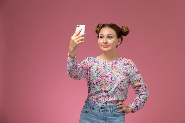 Vista frontale giovane femmina in fiore progettato camicia e blue jeans prendendo selfie su sfondo rosa