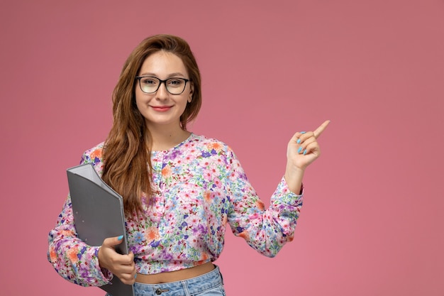 Vista frontale giovane femmina in fiore progettato camicia e blue jeans sorridente azienda documento grigio su sfondo rosa