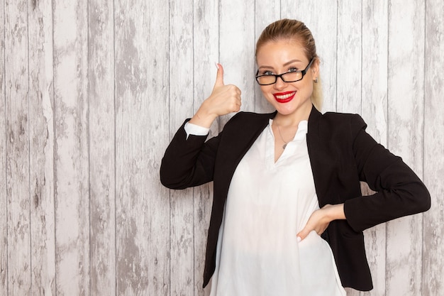 Vista frontale giovane imprenditrice in abiti rigorosi giacca nera con occhiali da sole ottici sorridente sulla parete bianca lavoro lavoro ufficio femmina riunione d'affari