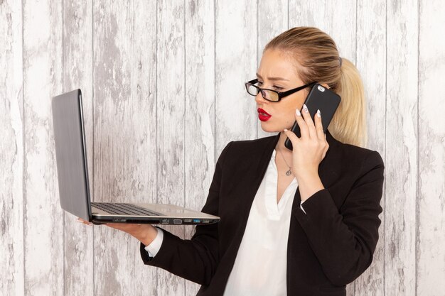 Vista frontale giovane imprenditrice in abiti rigorosi giacca nera utilizzando il suo computer portatile e parlando al telefono su superficie bianca