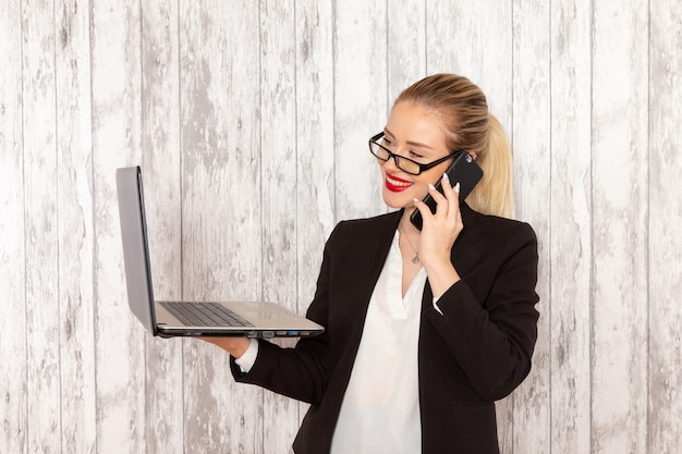 Vista frontale giovane imprenditrice in abiti rigorosi giacca nera utilizzando il suo computer portatile e parlando al telefono sulla scrivania bianca