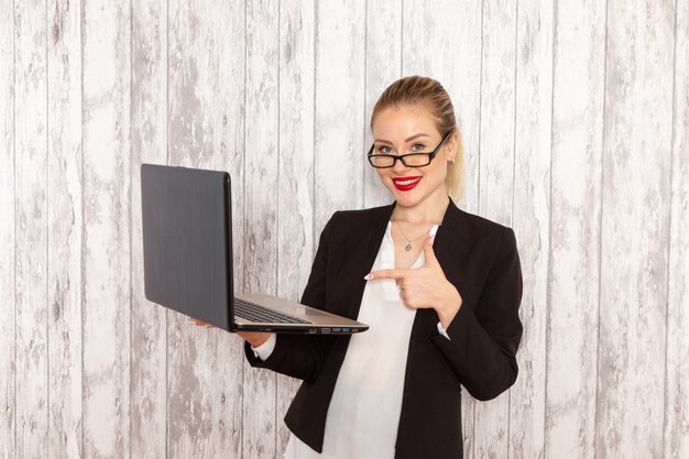 Vista frontale giovane imprenditrice in abiti rigorosi giacca nera utilizzando il suo computer portatile sullo scrittorio bianco chiaro