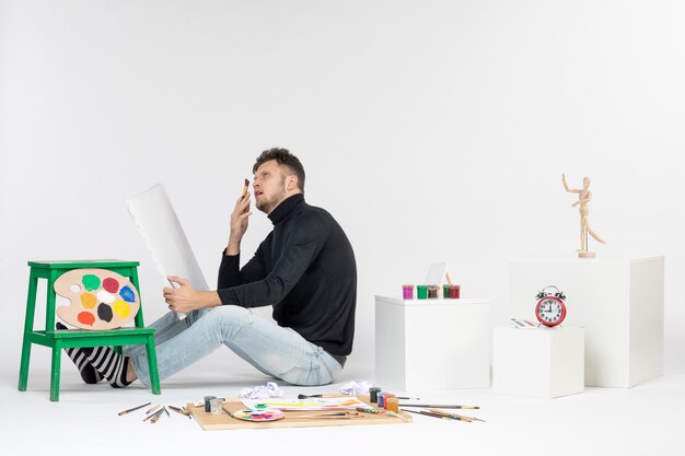 Vista frontale giovane maschio che prova a disegnare la pittura con la nappa sulle immagini della parete bianca dipinge l'artista del disegno della pittura di colore di arte