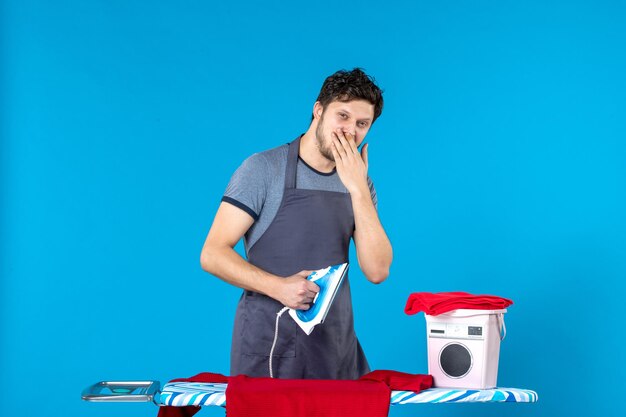 Vista frontale giovane maschio che stira la camicia rossa sulla superficie blu