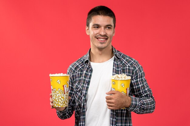 Vista frontale giovane maschio che tiene i pacchetti di popcorn con il sorriso sul film rosso chiaro del cinema del cinema della parete