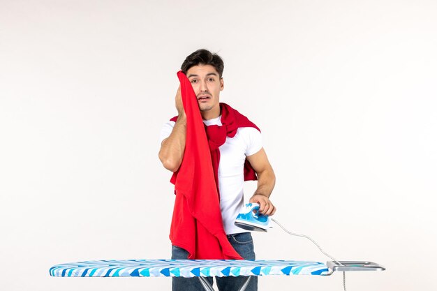 Vista frontale giovane maschio che tiene il ferro e l'asciugamano rosso sullo sfondo bianco colore della casa lavoro uomo lavori domestici bucato pulito emozione