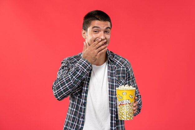 Vista frontale giovane maschio che tiene pacchetto di popcorn con risate sul film rosso chiaro del film del cinema del cinema della parete