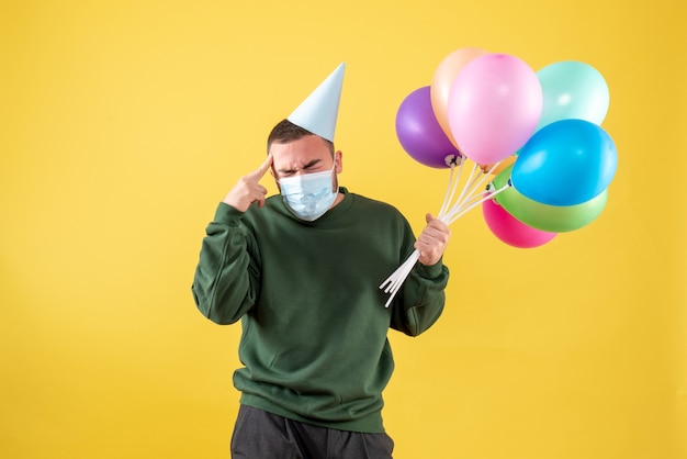 Vista frontale giovane maschio che tiene palloncini colorati in maschera su sfondo giallo