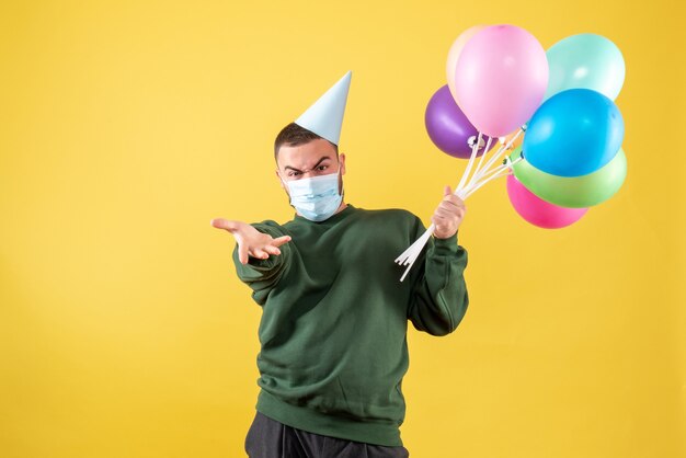 Vista frontale giovane maschio che tiene palloncini colorati in maschera su sfondo giallo