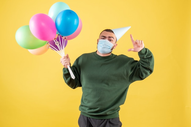 Vista frontale giovane maschio che tiene palloncini colorati su sfondo giallo