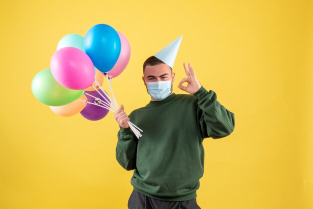 Vista frontale giovane maschio che tiene palloncini colorati su sfondo giallo