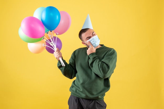 Vista frontale giovane maschio che tiene palloncini colorati su sfondo giallo