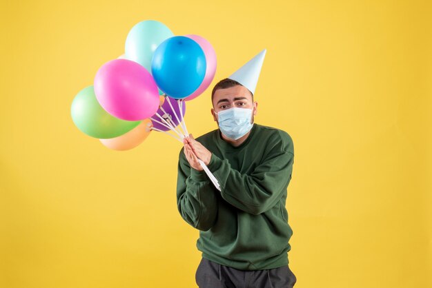 Vista frontale giovane maschio che tiene palloncini colorati su sfondo giallo