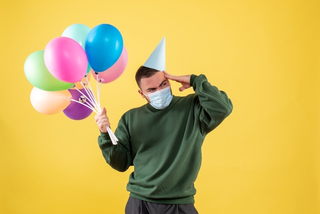 Vista frontale giovane maschio che tiene palloncini colorati su sfondo giallo