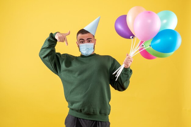 Vista frontale giovane maschio che tiene palloncini colorati su sfondo giallo