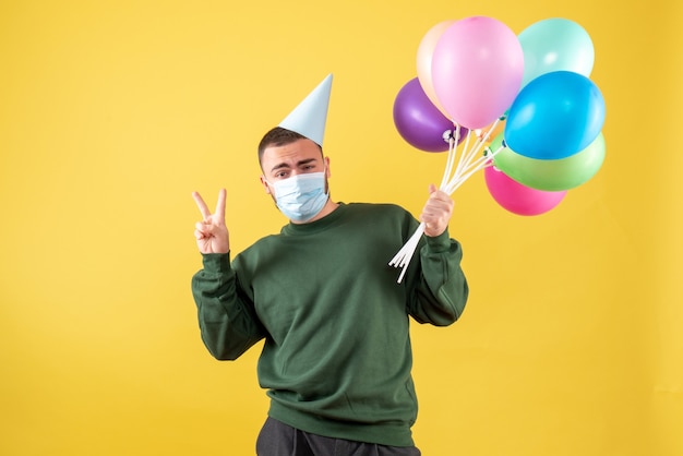 Vista frontale giovane maschio che tiene palloncini colorati su sfondo giallo
