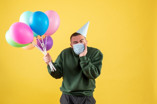 Vista frontale giovane maschio che tiene palloncini colorati su sfondo giallo