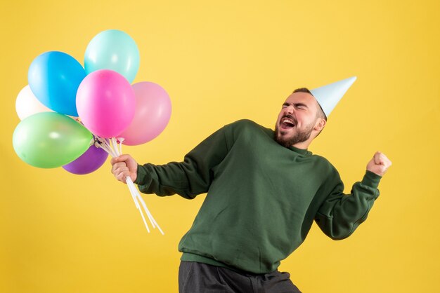 Vista frontale giovane maschio che tiene palloncini colorati su sfondo giallo