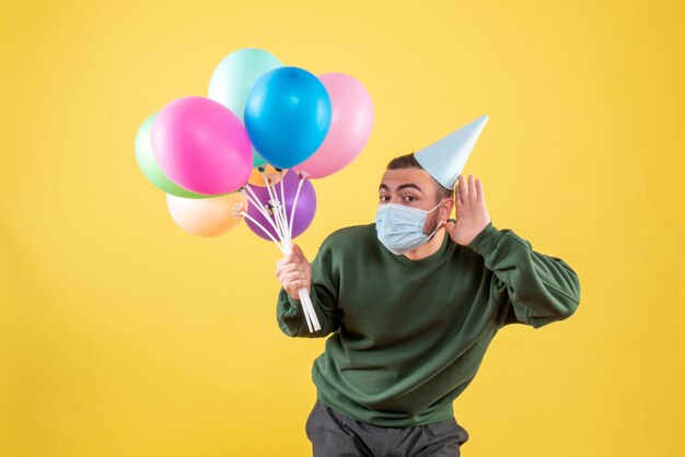 Vista frontale giovane maschio che tiene palloncini colorati su sfondo giallo