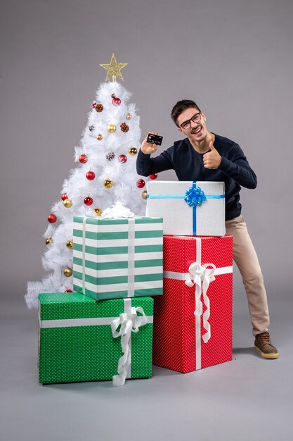 Vista frontale giovane maschio con carta di credito e regali su scrivania grigia vacanza capodanno natale