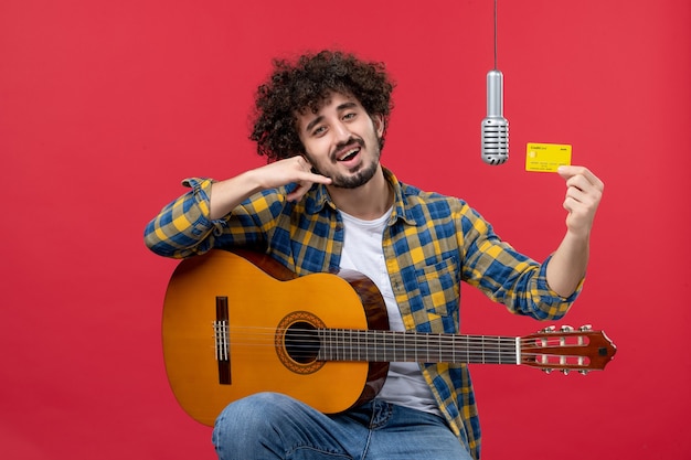 Vista frontale giovane maschio con la chitarra che tiene la carta di credito sul muro rosso performance dal vivo musicista concerto band denaro colore