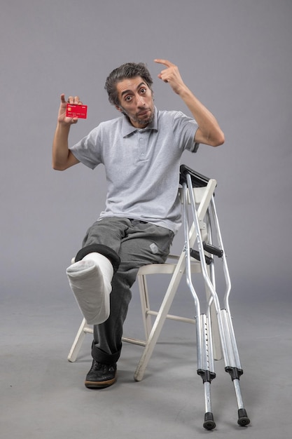Vista frontale giovane maschio con piede rotto e fasciatura in possesso di carta di credito sul muro grigio incidente torsione gamba piede dolore umano maschio