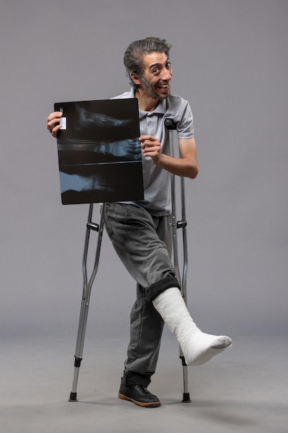 Vista frontale giovane maschio con piede rotto usando le stampelle e tenendo la sua radiografia sul muro grigio disabilita la torsione del piede del dolore da incidente rotto