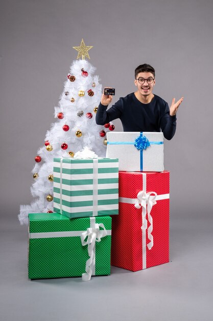 Vista frontale giovane maschio con regali e carta di credito sulla scrivania grigia vacanze di natale capodanno