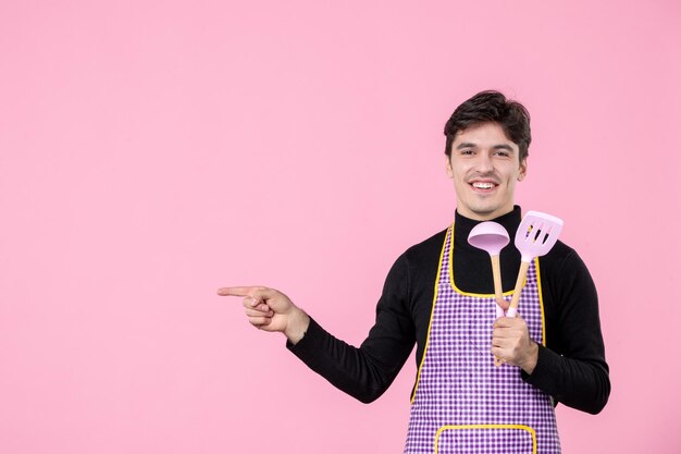 Vista frontale giovane maschio in mantello che tiene cucchiai rosa su sfondo rosa professione cibo cucina orizzontale lavoro colore cucina pasto pasta uniforme