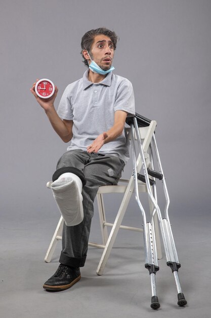 Vista frontale giovane maschio seduto con fasciatura legata a causa di un piede rotto e tenendo l'orologio sul muro grigio torsione dolore piede incidente gamba maschio
