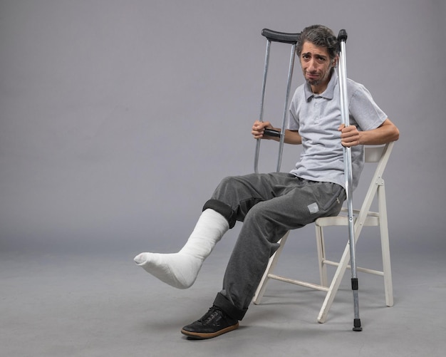 Vista frontale giovane maschio seduto con il piede rotto e tenendo le stampelle sul muro grigio piede dolore alla gamba rotta torsione incidente
