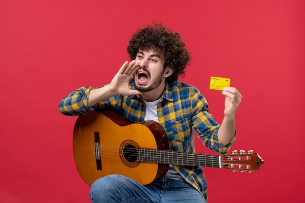 Vista frontale giovane maschio seduto con la chitarra in possesso di carta di credito sulla parete rossa colore performance concerto applauso musicista musica dal vivo