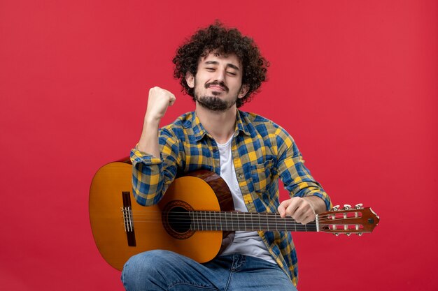 Vista frontale giovane maschio seduto con la chitarra sul muro rosso applauso musicista band suonare musica concerto dal vivo a colori