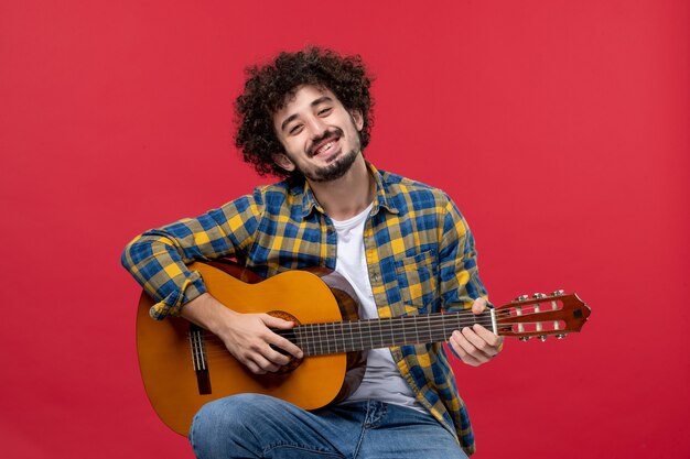 Vista frontale giovane maschio seduto con la chitarra sul muro rosso suona musica musicista colore applauso live