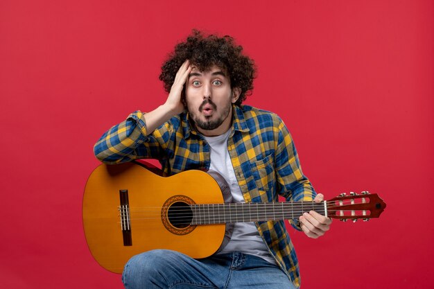 Vista frontale giovane maschio seduto con la chitarra sul muro rosso suona un concerto dal vivo musica applausi a colori