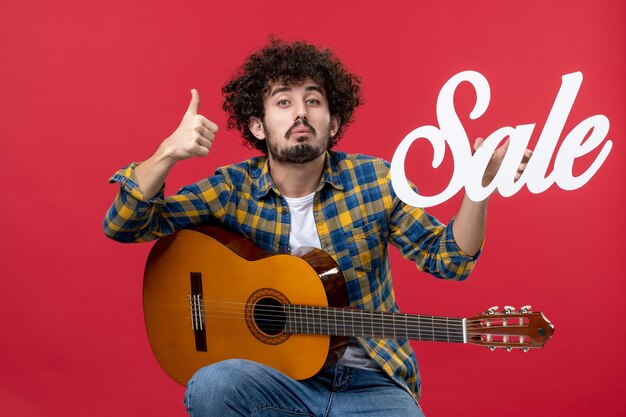 Vista frontale giovane maschio seduto con la chitarra sul muro rosso vendita di musica suona concerto applauso musicista dal vivo colore