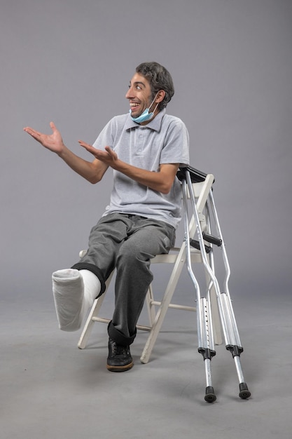 Vista frontale giovane maschio seduto con piede rotto bendato su scrivania grigia dolore torsione gamba incidente piede maschile