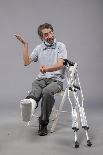 Vista frontale giovane maschio seduto con piede rotto e benda legata sul muro grigio dolore incidente piede torsione gamba maschio