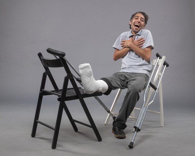 Vista frontale giovane maschio seduto con piede rotto e stampelle su scrivania grigia incidente torsione dolore alla gamba piede rotto