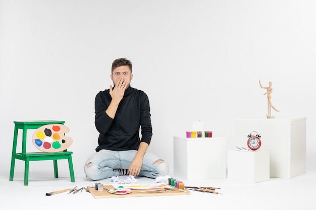 Vista frontale giovane maschio seduto intorno a pitture e nappe per disegnare sorpreso sul muro bianco artista di pittura di colore disegnare pittura d'arte