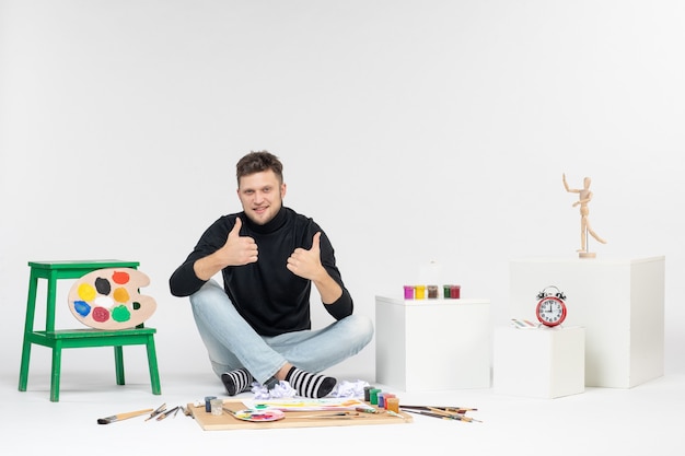 Vista frontale giovane maschio seduto intorno a vernici e nappe per disegnare su parete bianca arte disegnare pittura a colori artista pittura
