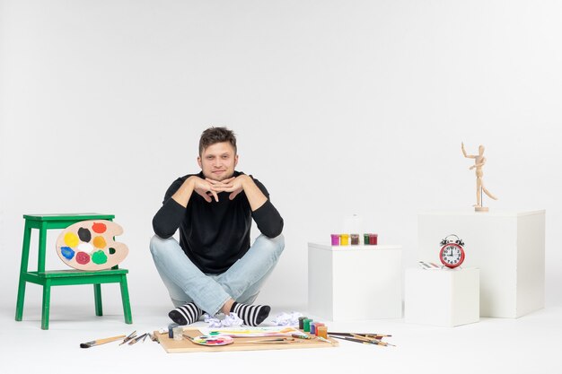 Vista frontale giovane maschio seduto intorno a vernici e nappe per disegnare sul muro bianco disegnare immagini a colori artista pittura pittura artistica
