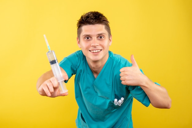Vista frontale giovane medico maschio in tuta medica tenendo l'iniezione su sfondo giallo