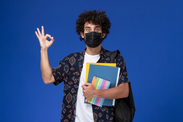 Vista frontale giovane studente maschio che indossa la maschera nera con zaino in possesso di file e quaderno su sfondo blu.