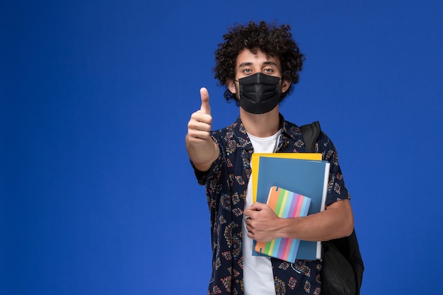 Vista frontale giovane studente maschio che indossa la maschera nera con zaino in possesso di quaderno e file su sfondo blu.