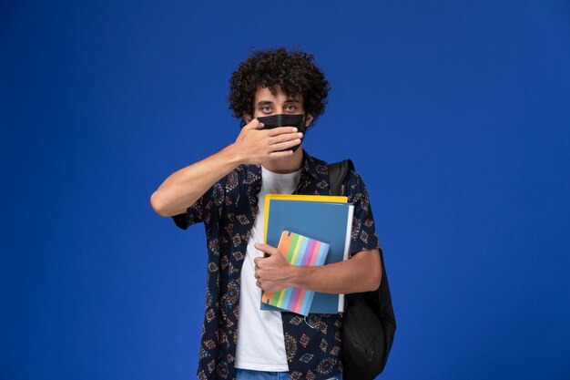 Vista frontale giovane studente maschio che indossa la maschera nera con zaino in possesso di quaderno e file sullo scrittorio blu.